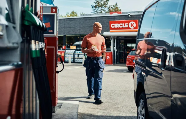 Man returning to his vehicle at a Circle K station with a sandwich and coffee in hand